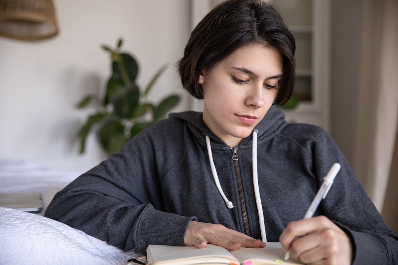 Young woman in a hoodie writing in a notebook while sitting on a bed indoors.