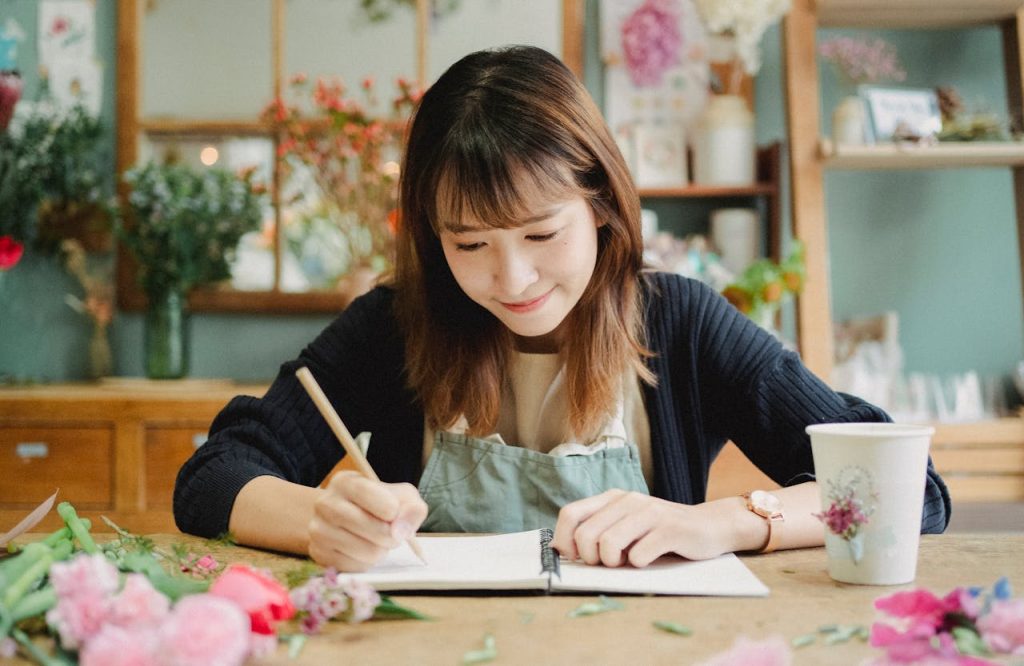 Glad young Asian female in uniform taking notes in notebook while working in light floral shop and sitting at table covered with many flowers petals
