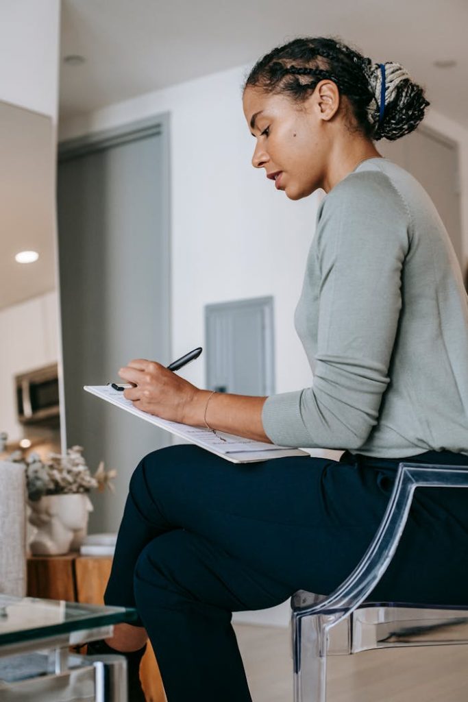 A young woman writes in a notebook while seated in a modern living room, focused and calm.