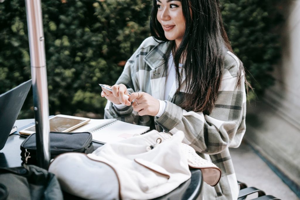 Positive young Asian female student in checkered shirt sitting at table in campus and taking notes while doing home task