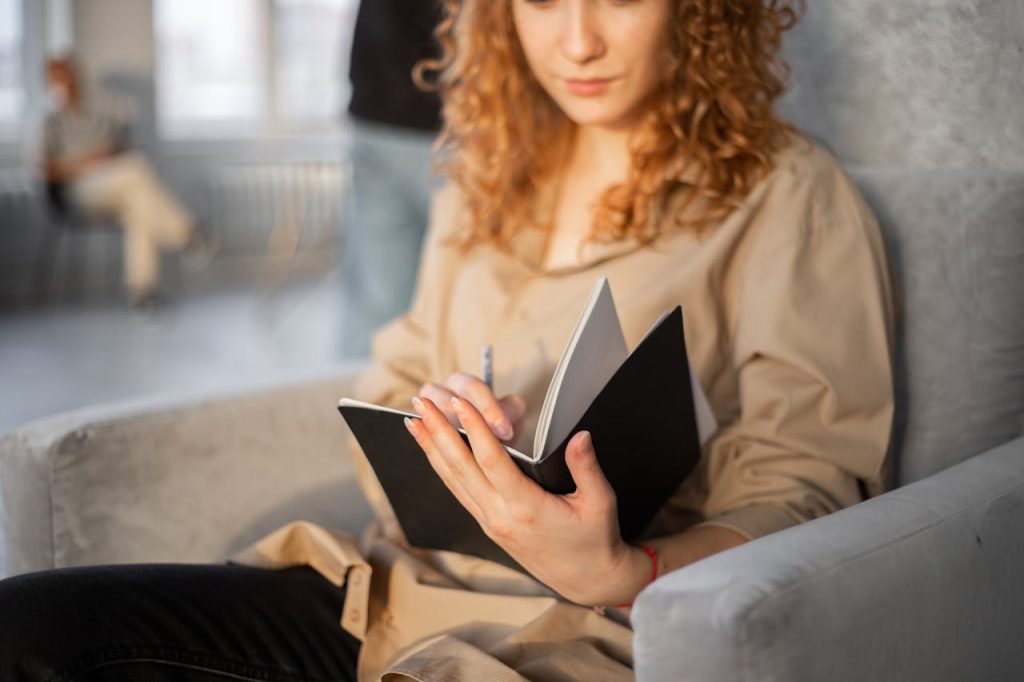 Young crop anonymous female with pen taking notes in planner while resting in soft gray armchair on blurred background